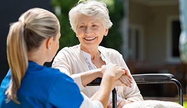 Smiling senior patient sitting on wheelchair with nurse supporting her. Doctor looking at elderly patient on a wheelchair in the garden. Nurse holding hand of mature woman outside pension home.