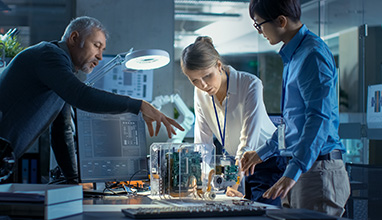 Team of Computer Engineers Lean on the Desk and Choose Printed Circuit Boards to Work with, Computer Shows Programming in Progress. In The Background Technologically Advanced Scientific Research Center.