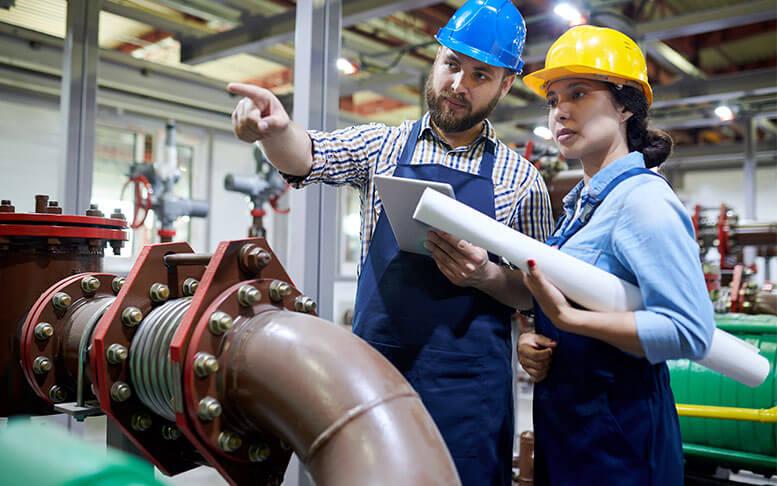 Portrait of two factory workers pointing away while working with piping and machines in modern workshop.