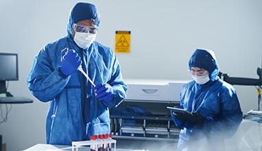 Serious concentrated African-American virologist in protection workwear standing at table and dropping blood in test tube while Asian colleague examining information on tablet