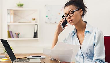 Young serious african-american businesswoman talking by phone with papers, sitting at modern office workplace. Business consulting, copy space