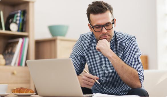Man sitting in his living room with his laptop open and pen in hand looking thoughtfully at his cover letter.